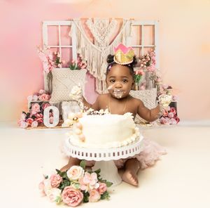 Decorative party setup with macrame backdrop, pink roses and white cake on stand against pink background.