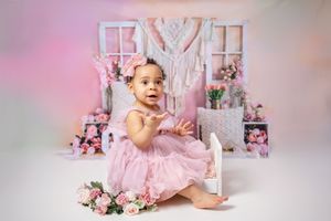 A joyful toddler in a pink tulle dress sits in front of a floral backdrop with pink decorations and white window frames.