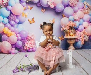 A baby sits in a dreamy pastel pink and purple first birthday setup with balloon arch and butterfly decorations.