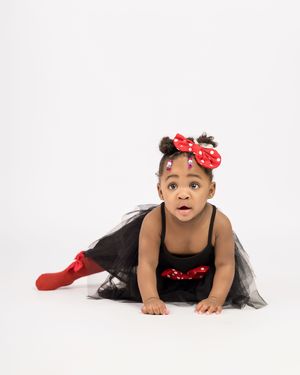 Young dancer in a black tutu and red accessories poses on the floor against a white backdrop.