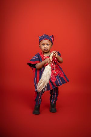 Small child in traditional Nigerian attire with blue and navy striped agbada poses against a red backdrop.