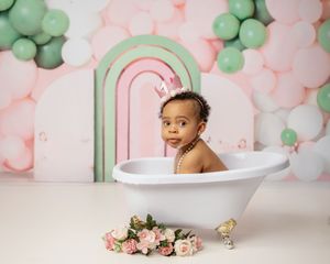 Baby sitting in a decorative bathtub with pink and mint green balloon arch and floral decorations for milestone photo session.