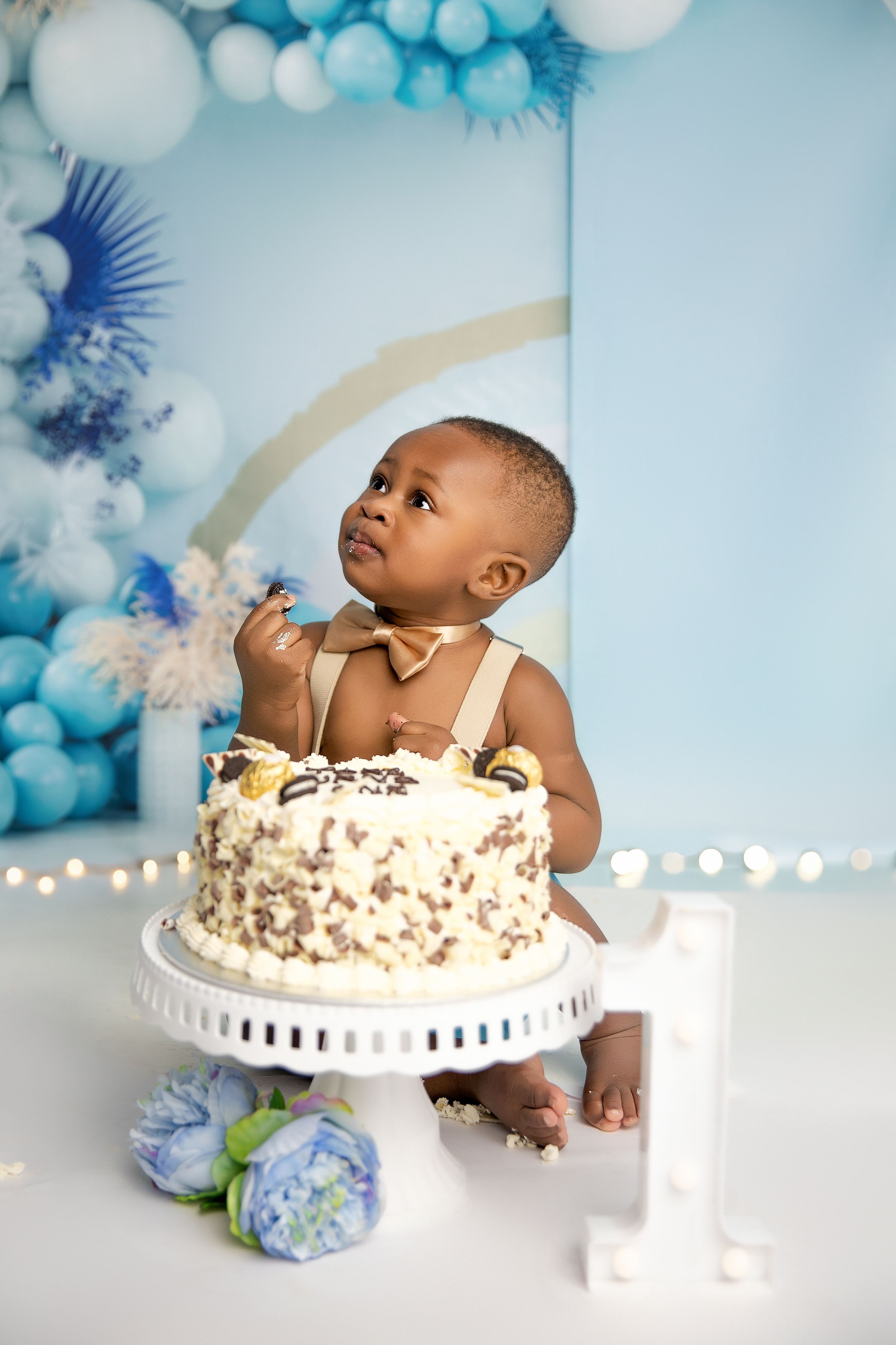 Baby sits near blue balloon arch and white cake stand during first birthday photo shoot with white and blue decor.