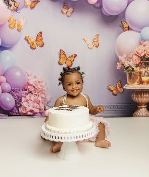 Festive butterfly-themed birthday setup with purple balloons, floral decorations and white tiered cake on a pedestal.