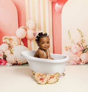 Baby sits in decorative white bathtub surrounded by pink balloons and floral arrangements for milestone photo shoot.