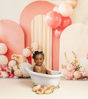 Pink balloon arch and floral decor frames a bathtub setup for a baby photography session with soft pastel colors.