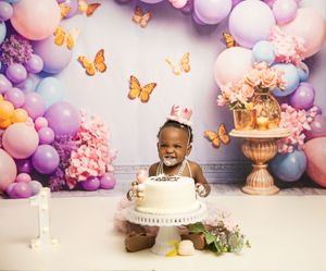 A cake smash photo setup with purple and pink balloon arch, orange butterflies, and floral decor against a white backdrop.