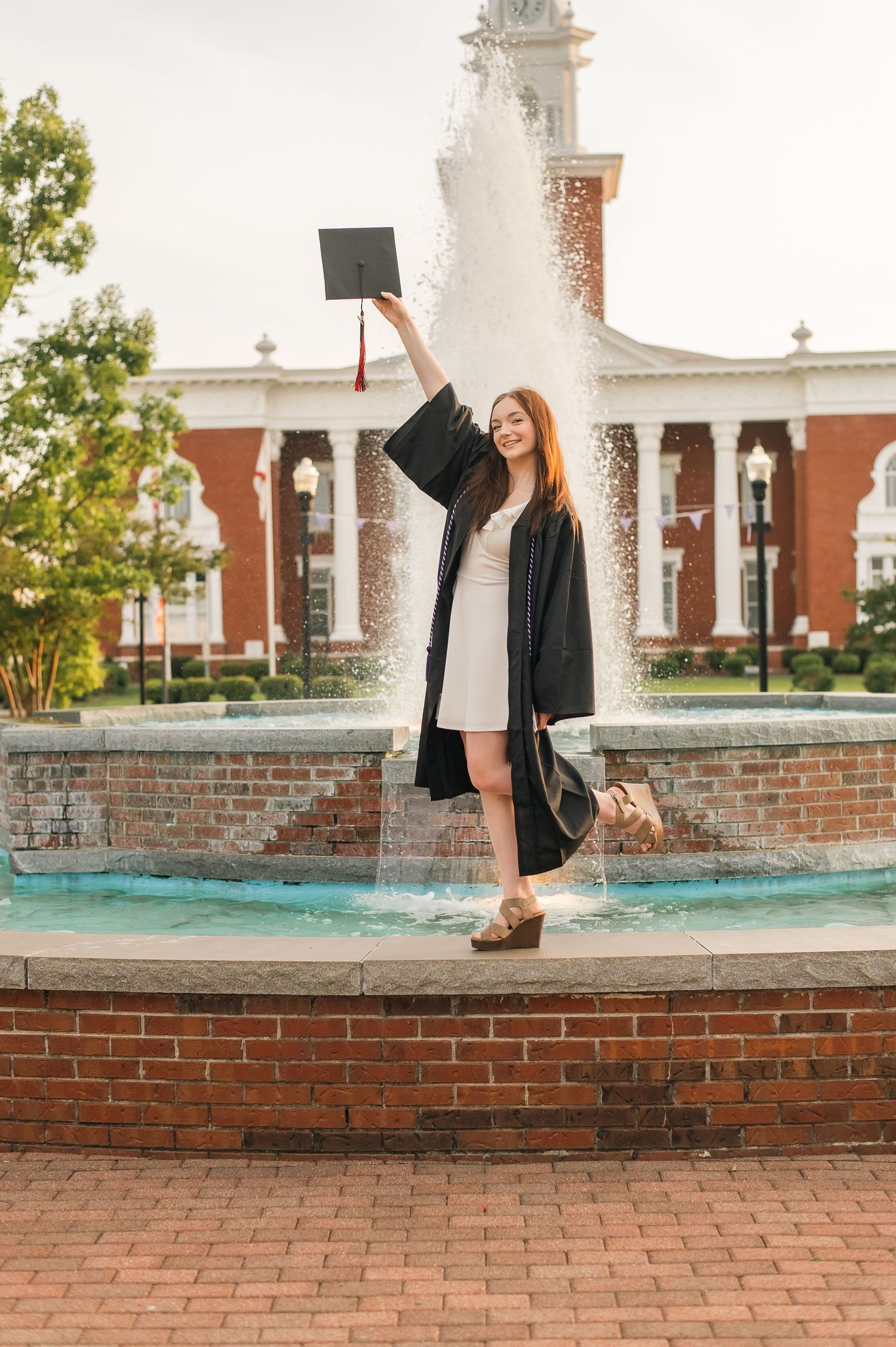 Graduate celebrates with cap raised by fountain outside brick university building with white columns and clocktower.