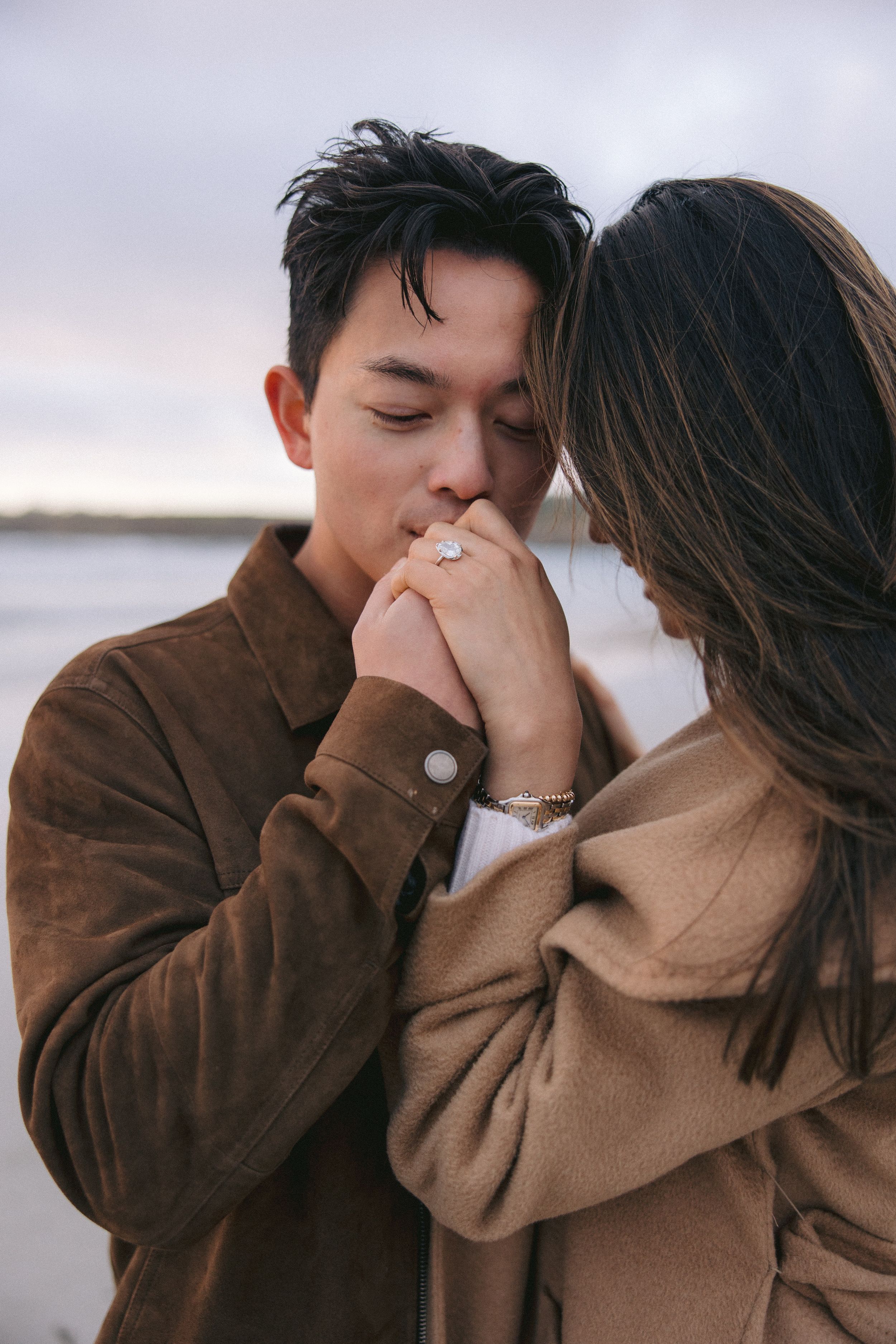 Romantic couple in brown coats sharing an intimate moment by the water at sunset.
