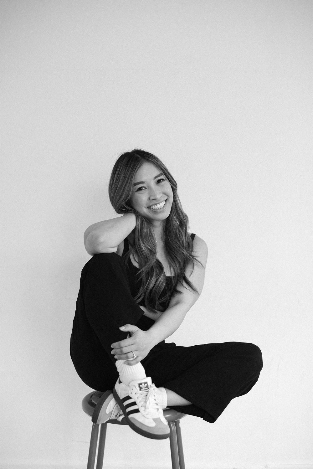 Person in black attire poses playfully on a stool against minimalist white background.