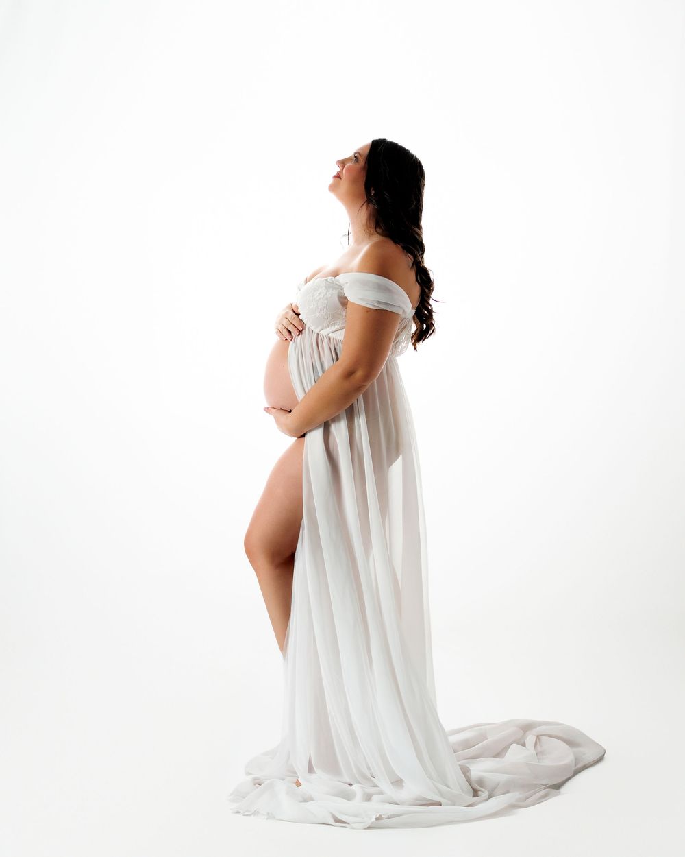 Person in white flowing dress poses gracefully against white background in artistic maternity photoshoot.