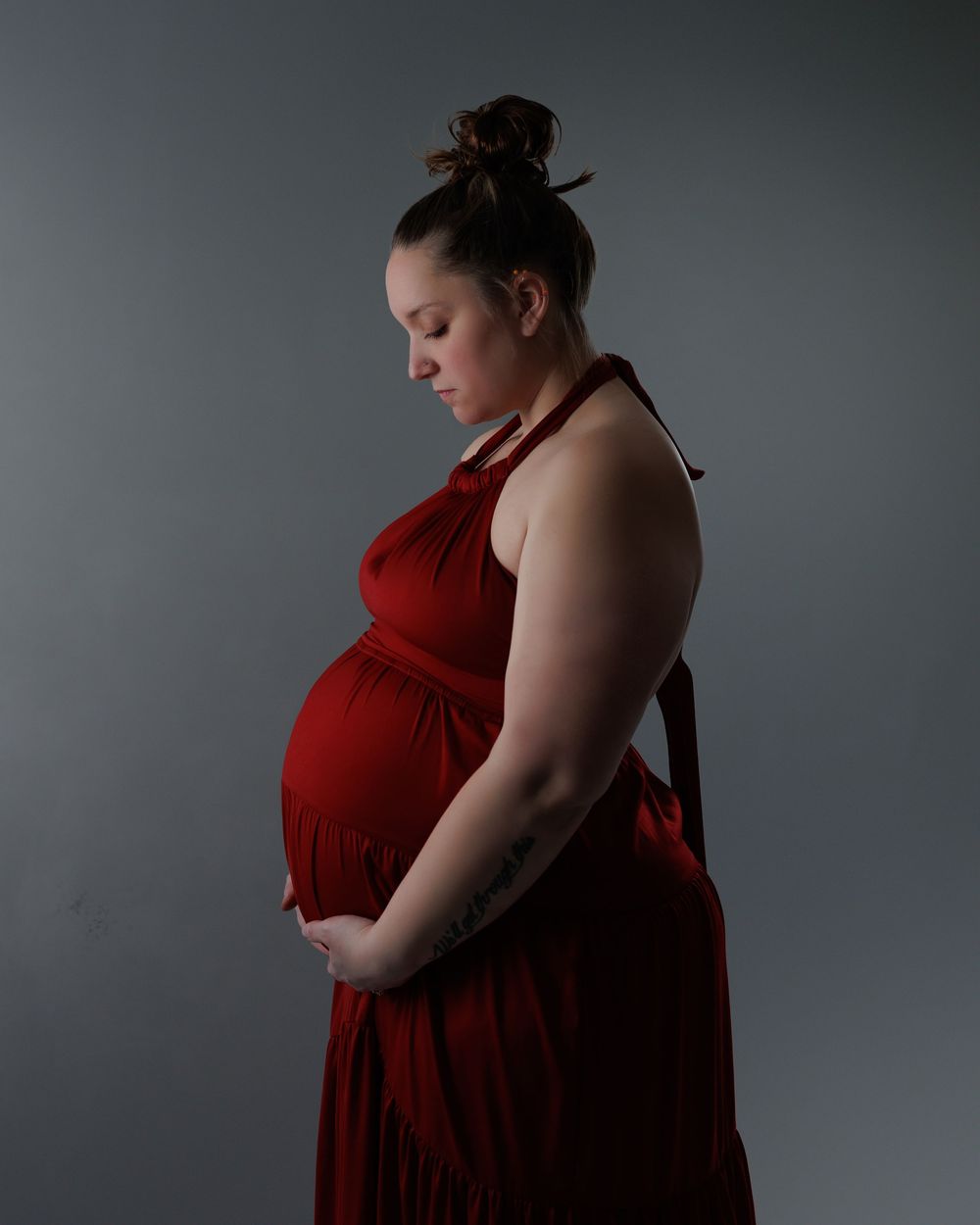 A person in a flowing red dress poses against a gray background in a dramatic maternity portrait.