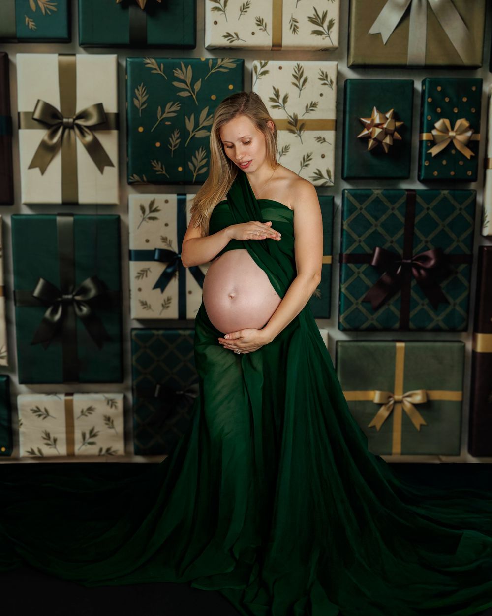Elegant maternity photo featuring an emerald green flowing gown against a backdrop of decorative gift boxes and bows.
