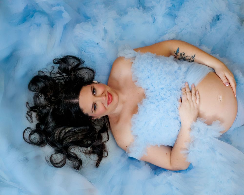 A woman with dark hair lies in a milky blue bath with scattered flower petals creating a dreamy spa-like atmosphere.