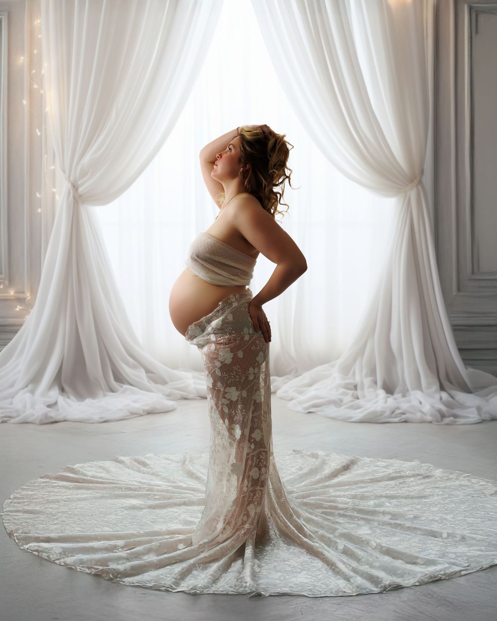 An elegant maternity photo featuring a sheer lace gown and flowing white curtains in a bright studio setting.
