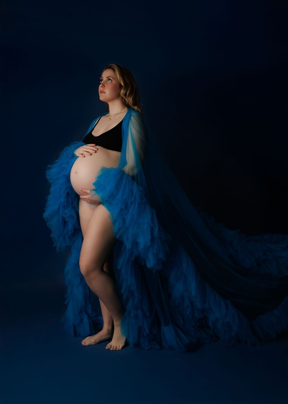 Artistic maternity photo against dark blue background with flowing blue sheer fabric draping around expectant mother.