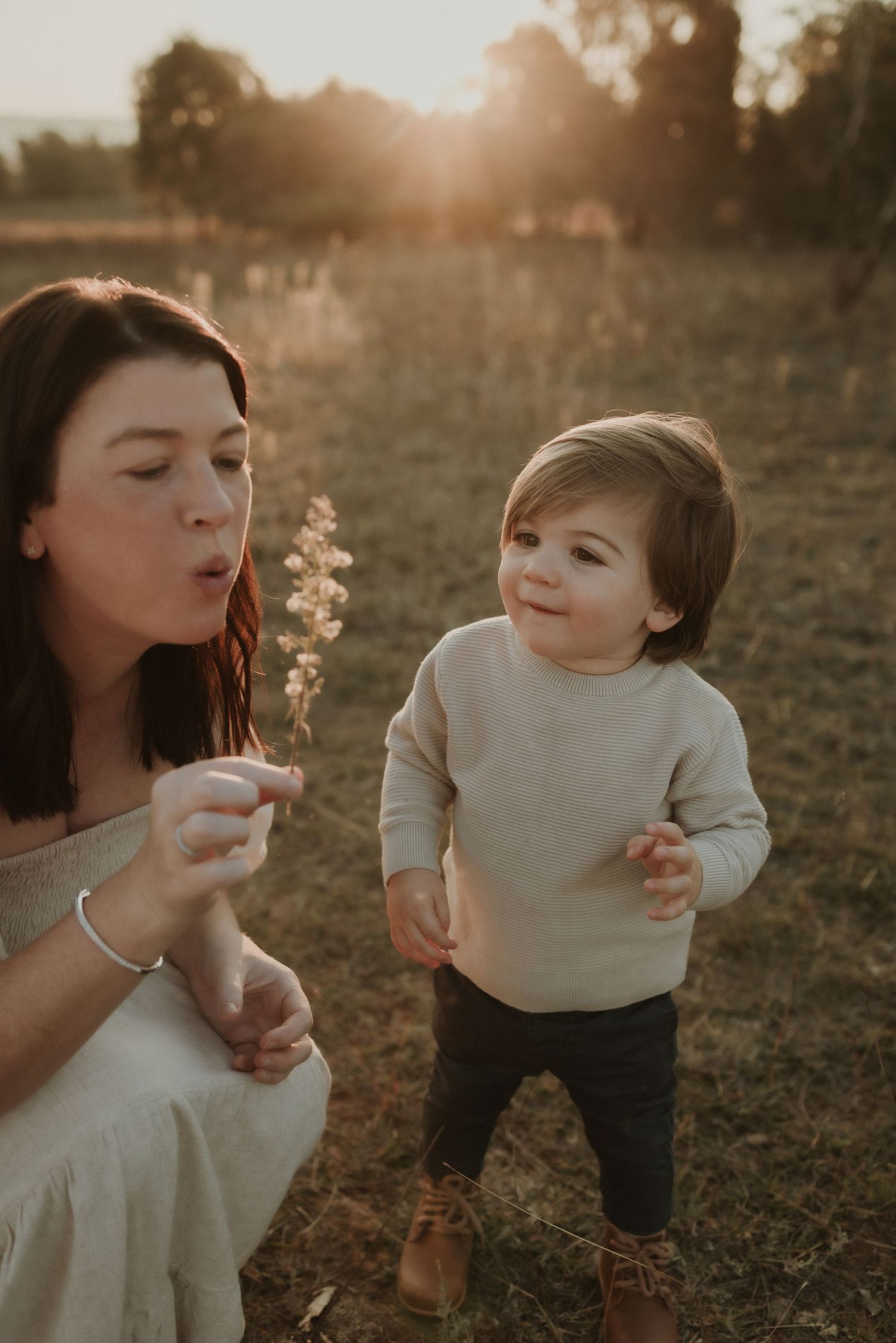 A young child with his mother blowing dandelion seeds in a sunlit Queanbeyan field at golden hour.