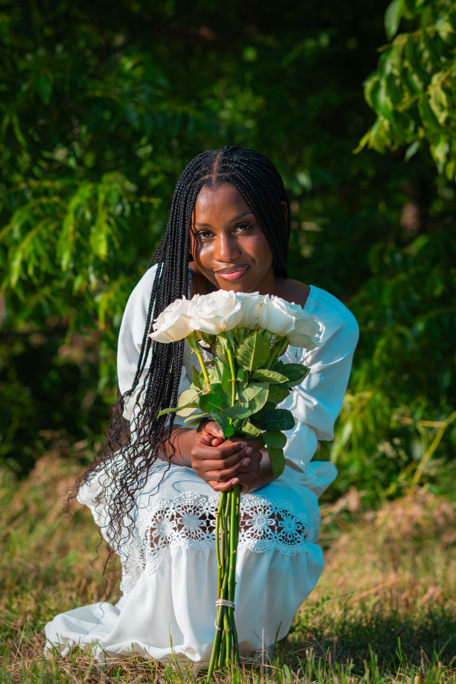 A person in a white dress kneels on grass holding white roses against a backdrop of lush green foliage.