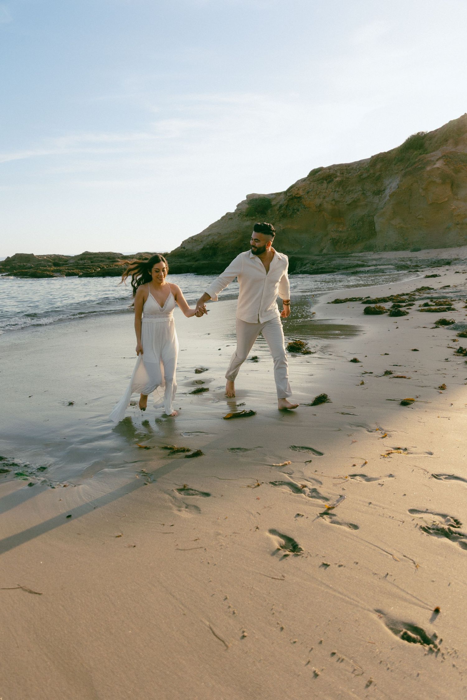 Two people in white outfits walking hand in hand along a scenic beach with cliffs during a sunny afternoon.