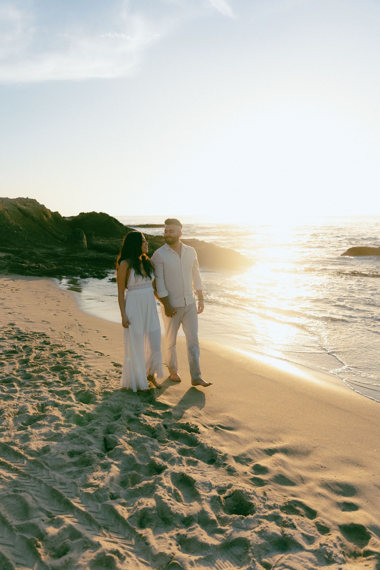 A couple in white outfits walking along a beach at sunset with waves crashing on rocky shore in the background.