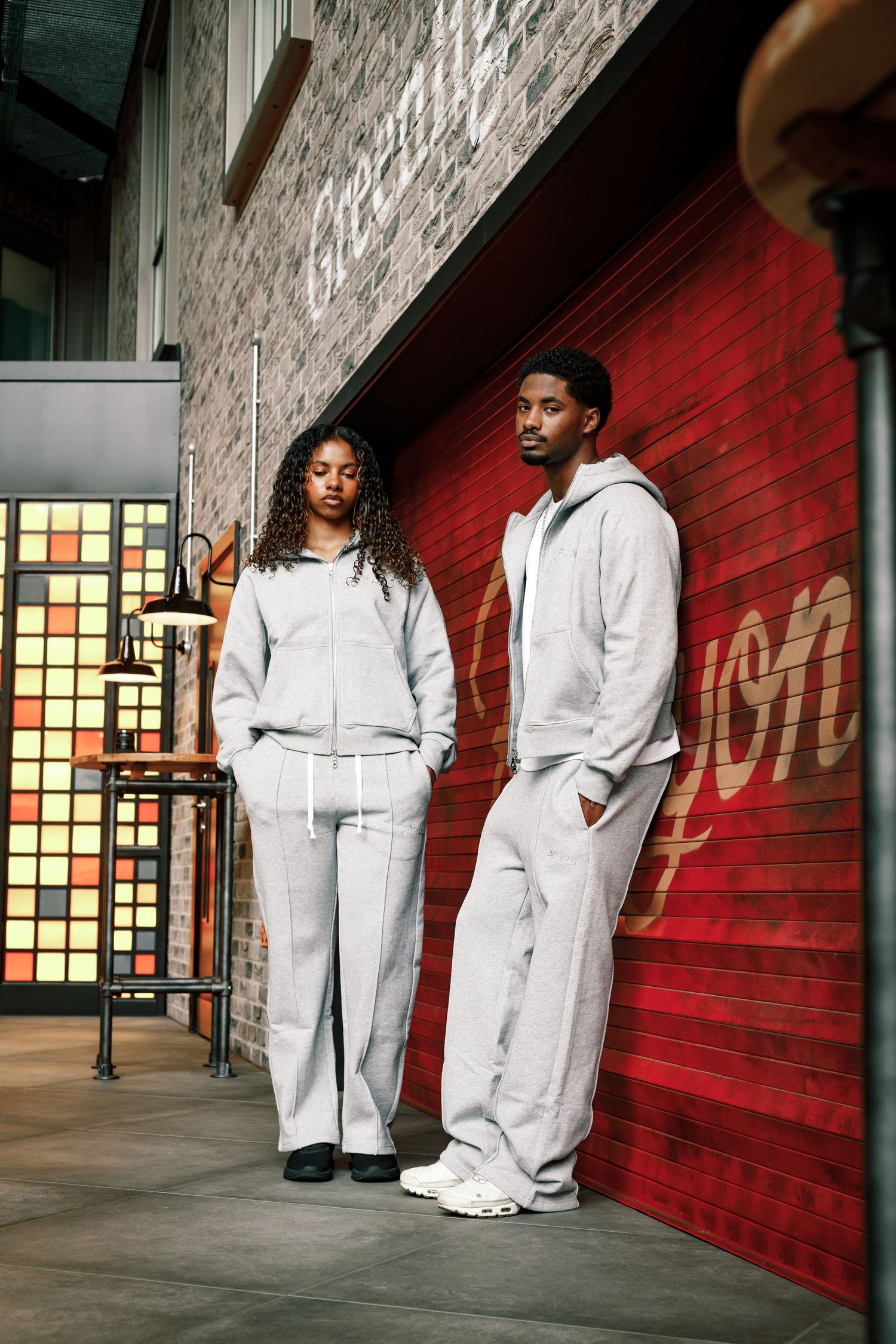Two people wearing casual loungewear, standing back to back against gray background, with gray sweatpants and pink/white tops.