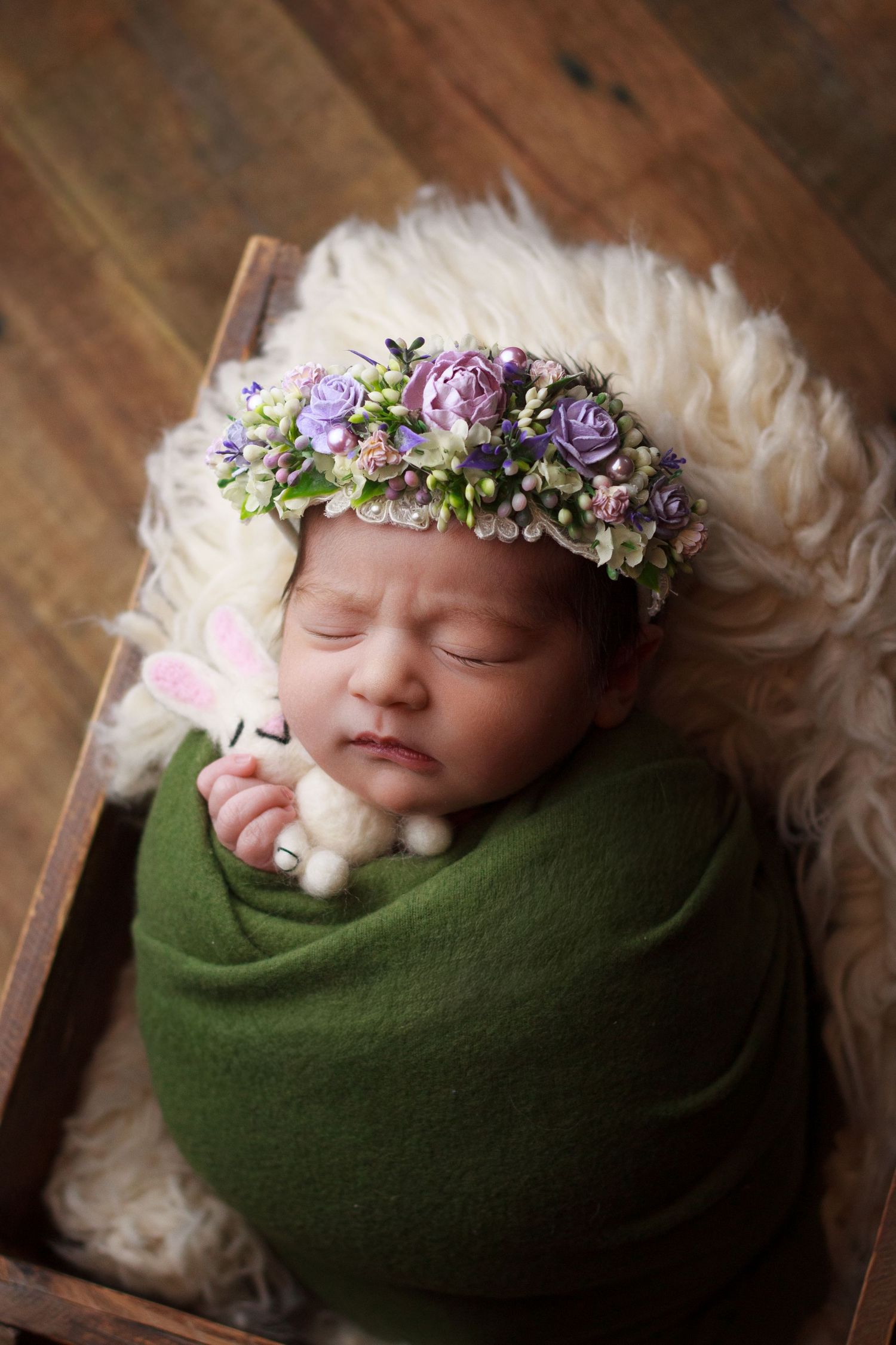 Newborn baby girl wearing a purple and green floral crown and wrapped in dark olive green wrap shown sleeping in a wooden box