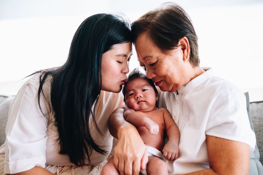 Mother and grandmother share a moment with a newborn baby between them in a warm indoor setting taken by Melbourne newborn photographer