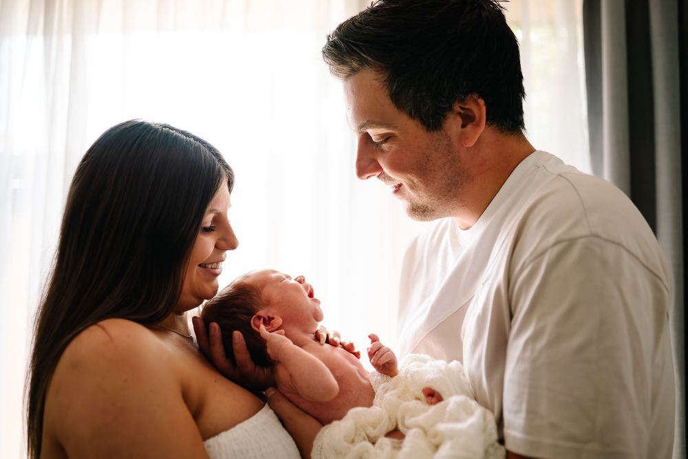 Parents share an intimate moment with their newborn baby beside a bright window with white curtains taken by Melbourne newborn photographer.