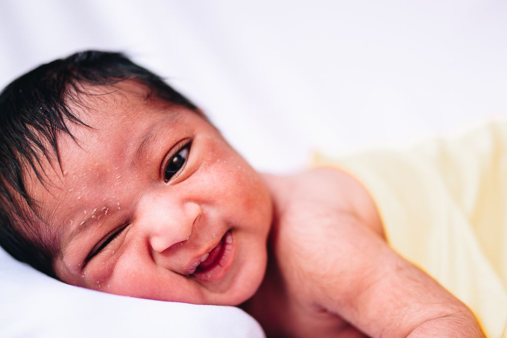 A happy newborn smiling while lying on a white sheet with yellow blanket taken by Melbourne newborn photographer.