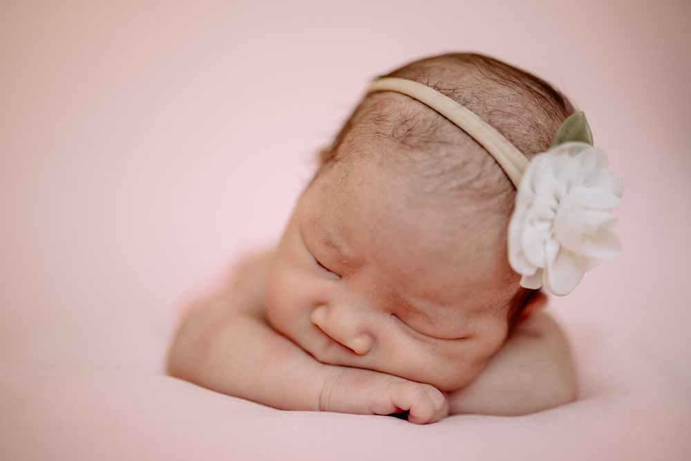 A sleeping newborn with a delicate white flower headband rests against a soft pink background taken by Melbourne newborn photographer.