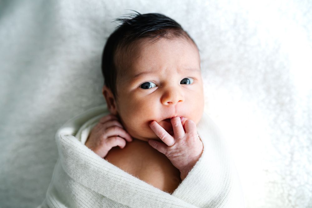 A newborn baby smiles while wrapped in a soft white blanket against a light background taken by Melbourne newborn photographer.