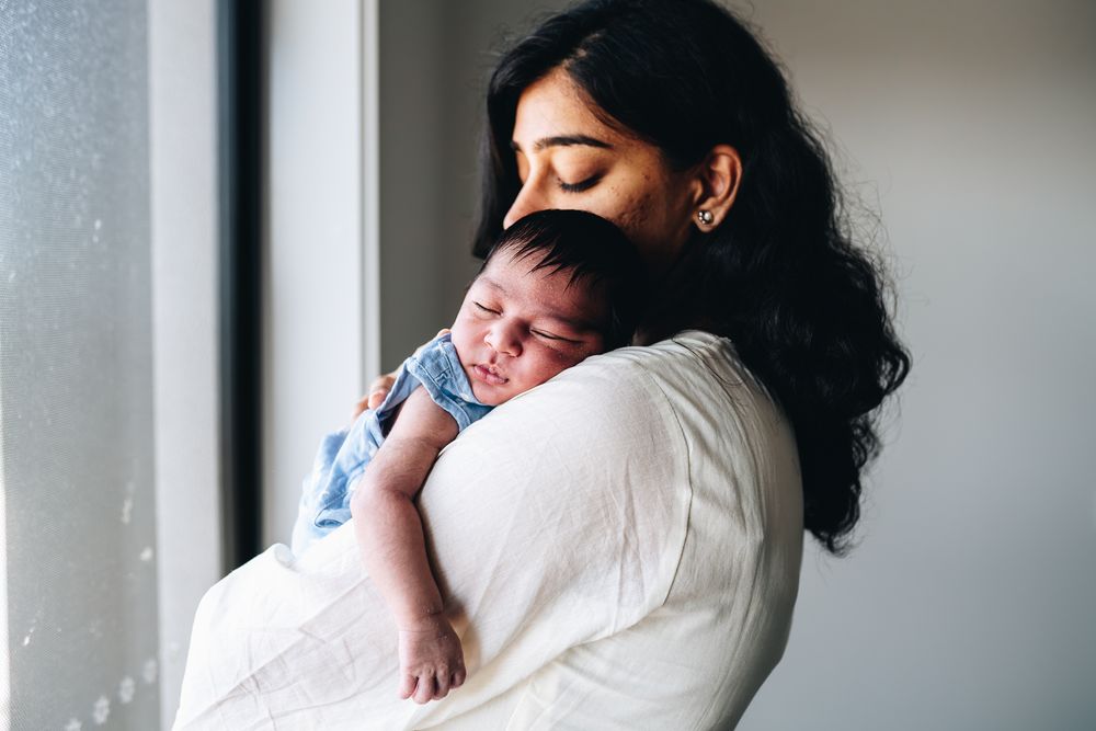 A woman in a white dress holds a sleeping baby near a window while standing in soft natural light taken by Melbourne newborn photographer.
