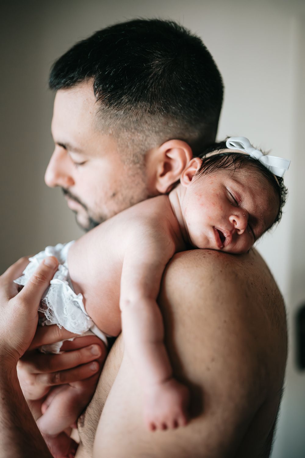 A tender moment captured as a newborn baby in a white bow sleeps peacefully on a father's shoulder taken by Melbourne newborn photographer.