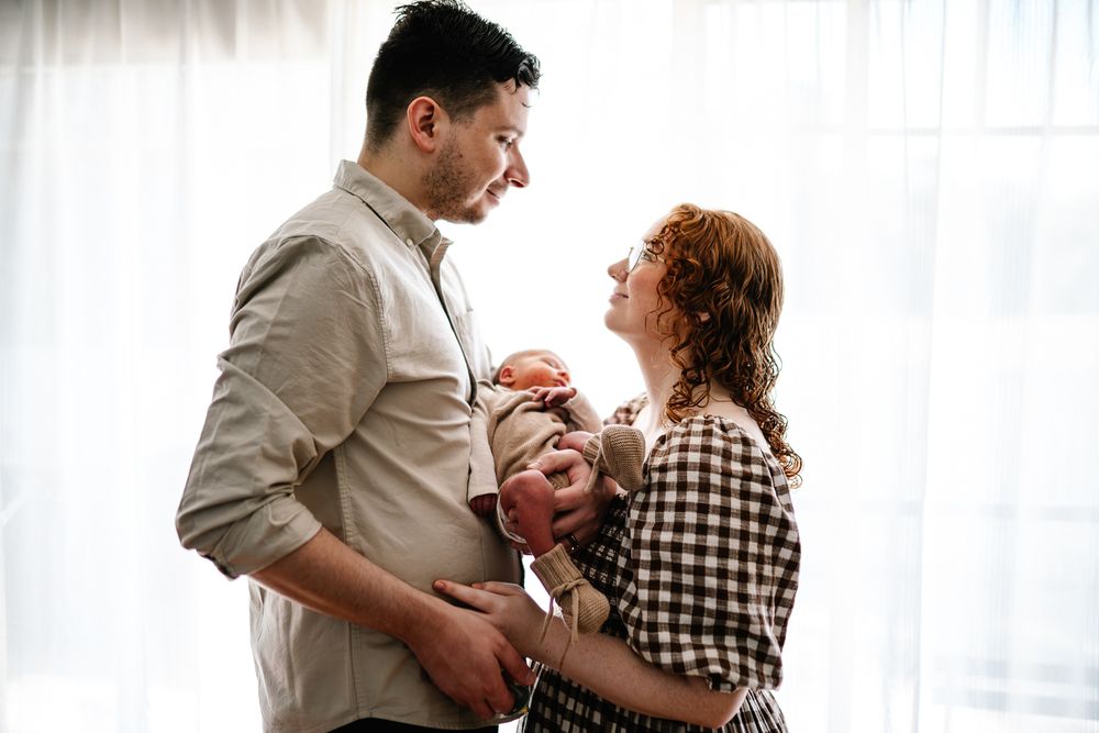A couple holds and comforts their newborn baby together in front of bright curtained windows.