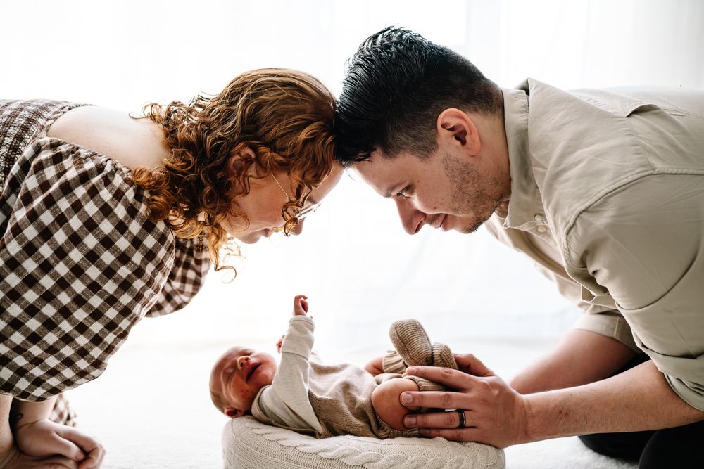 Parents lovingly lean over their happy baby lying on a white surface in a bright natural light setting.