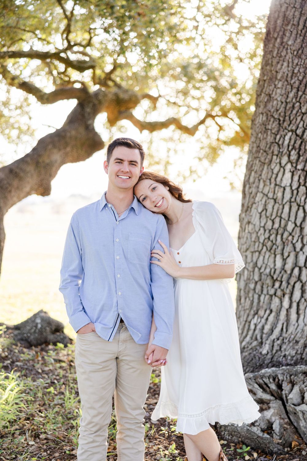 A couple poses casually together under a large oak tree in soft natural lighting.