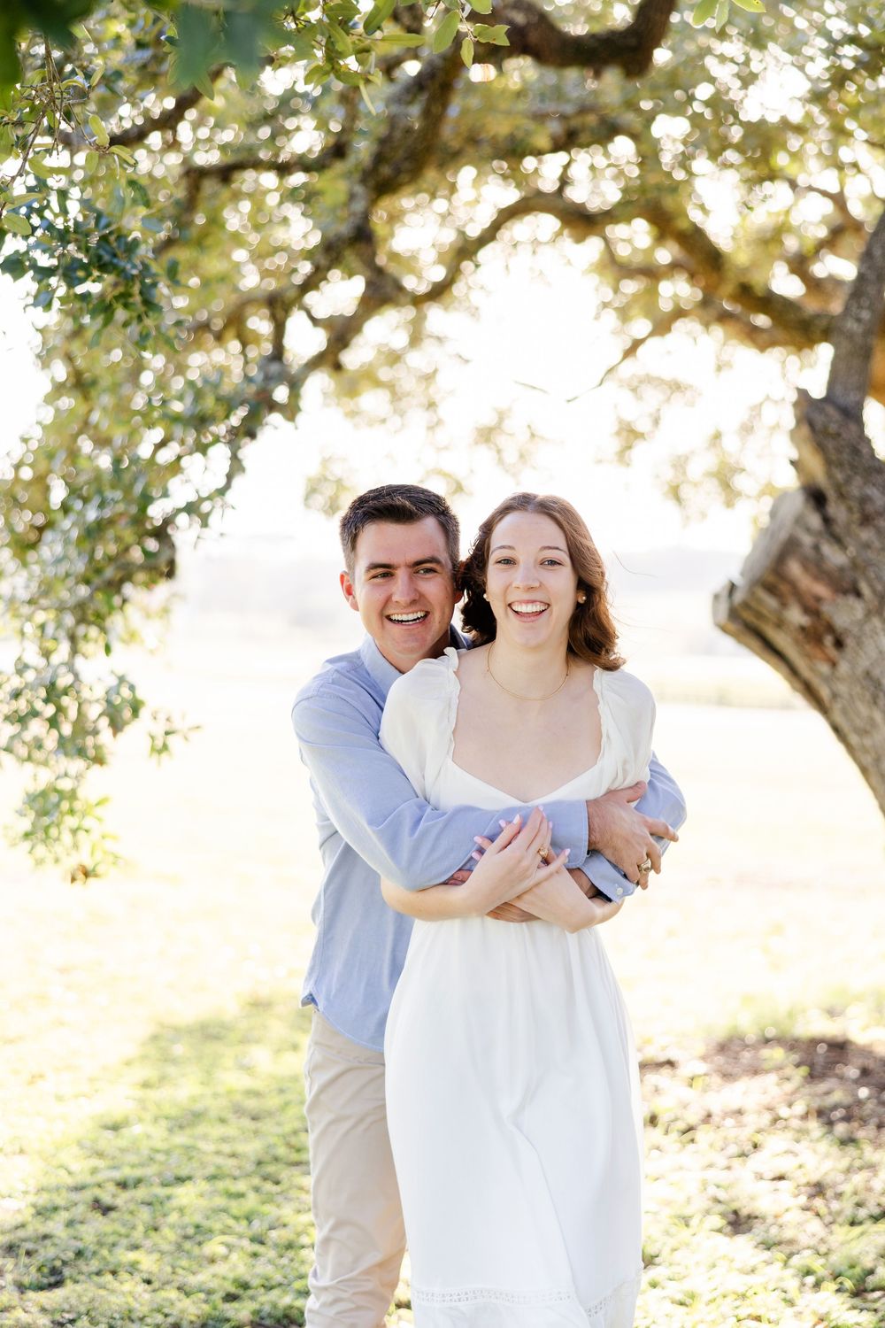 A couple embraces and laughs together under a sunlit tree in a dreamy outdoor setting.