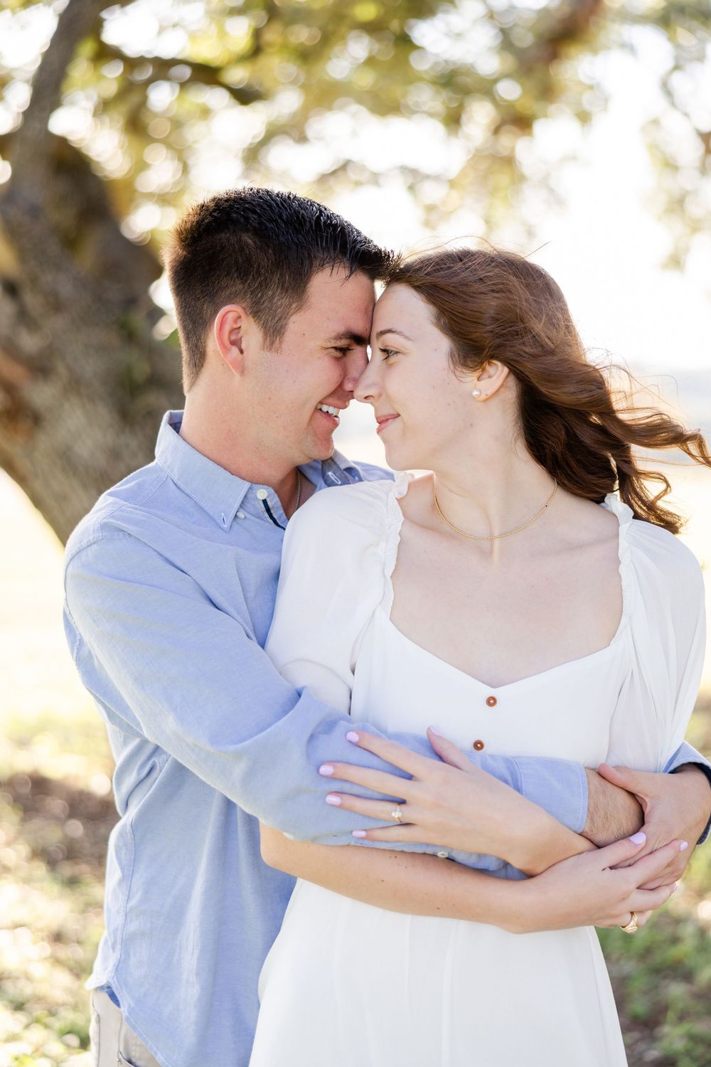 A romantic outdoor couples portrait with soft natural lighting shows two people embracing and smiling in a tender moment.