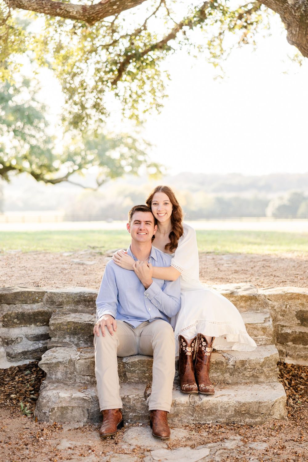 A couple sits on stone steps under a sunlit tree, wearing casual attire with brown boots and a white dress.