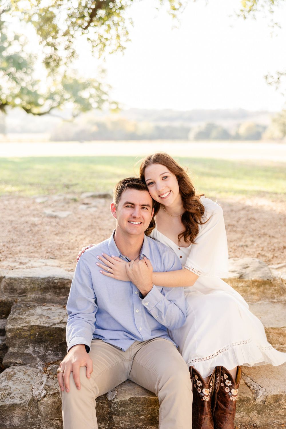 Romantic couple sitting together on stone wall during golden hour engagement photoshoot in rustic outdoor setting.