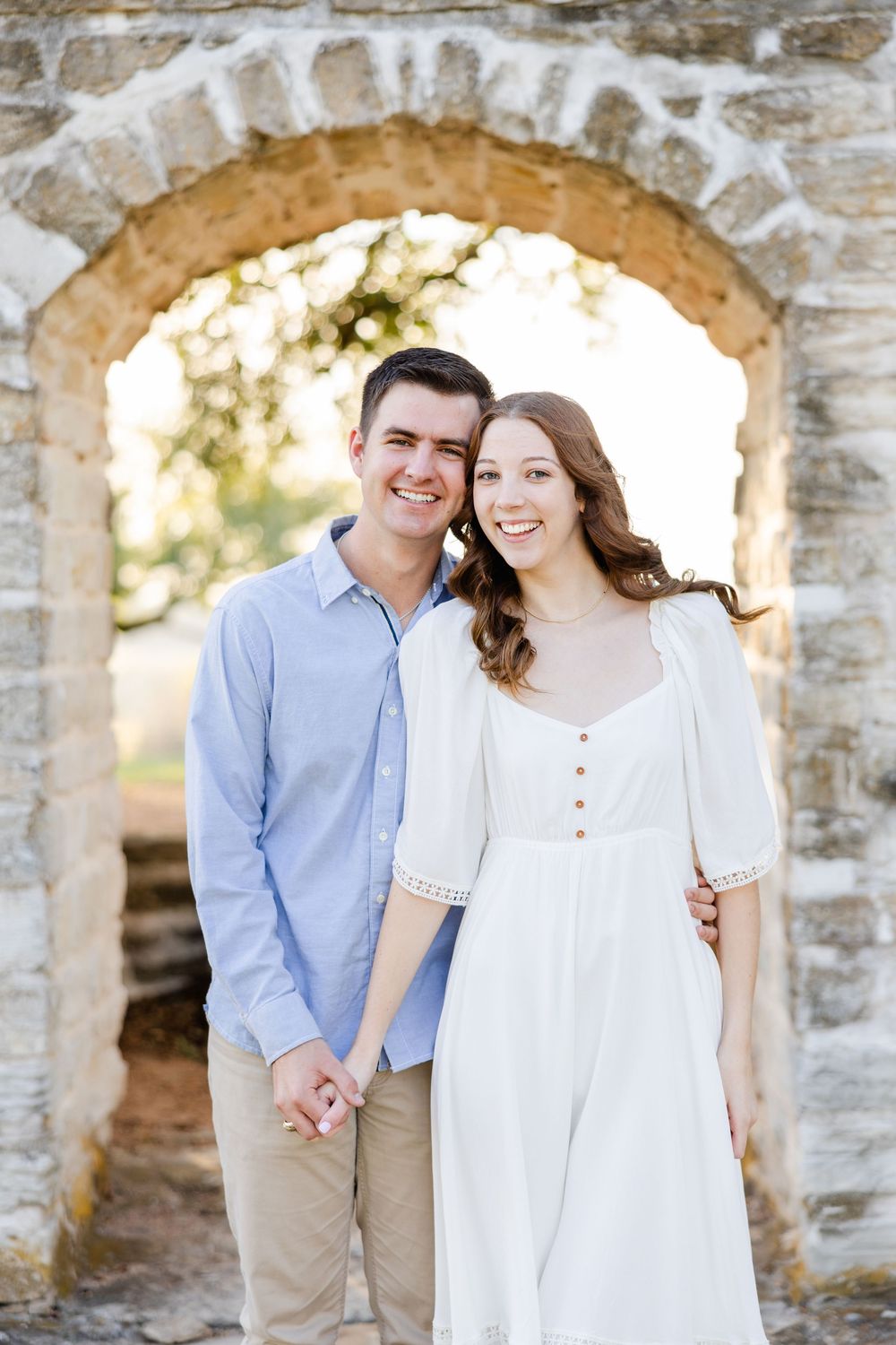 A happy couple poses under a stone archway, wearing coordinated outfits of blue and white on a sunny day.