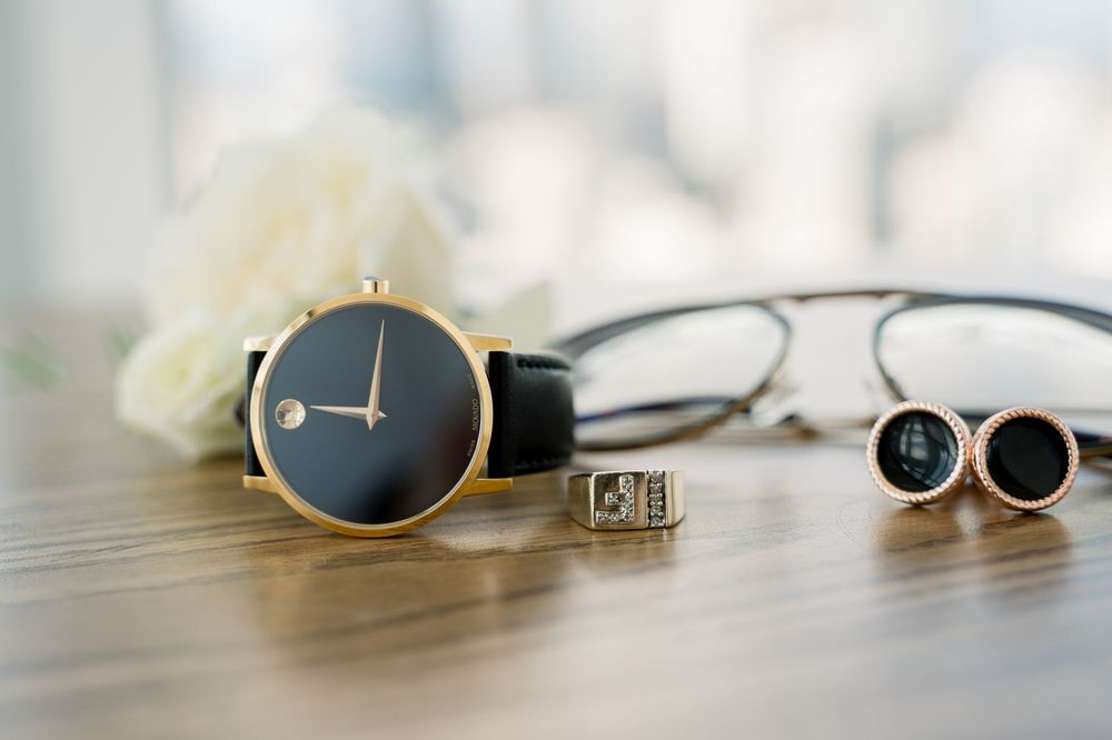 Minimalist black and gold watch displayed with eyeglasses and cufflinks on wooden surface in soft lighting.