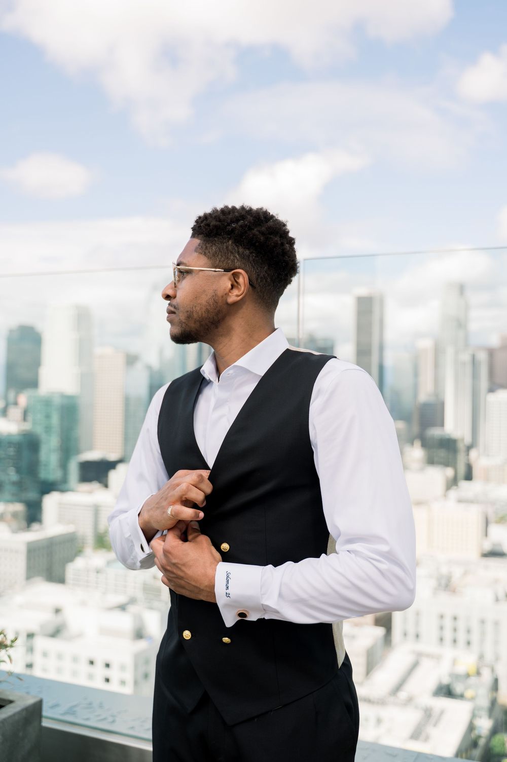Professional businessman in formal attire adjusts sleeve while standing on rooftop with urban skyline backdrop.