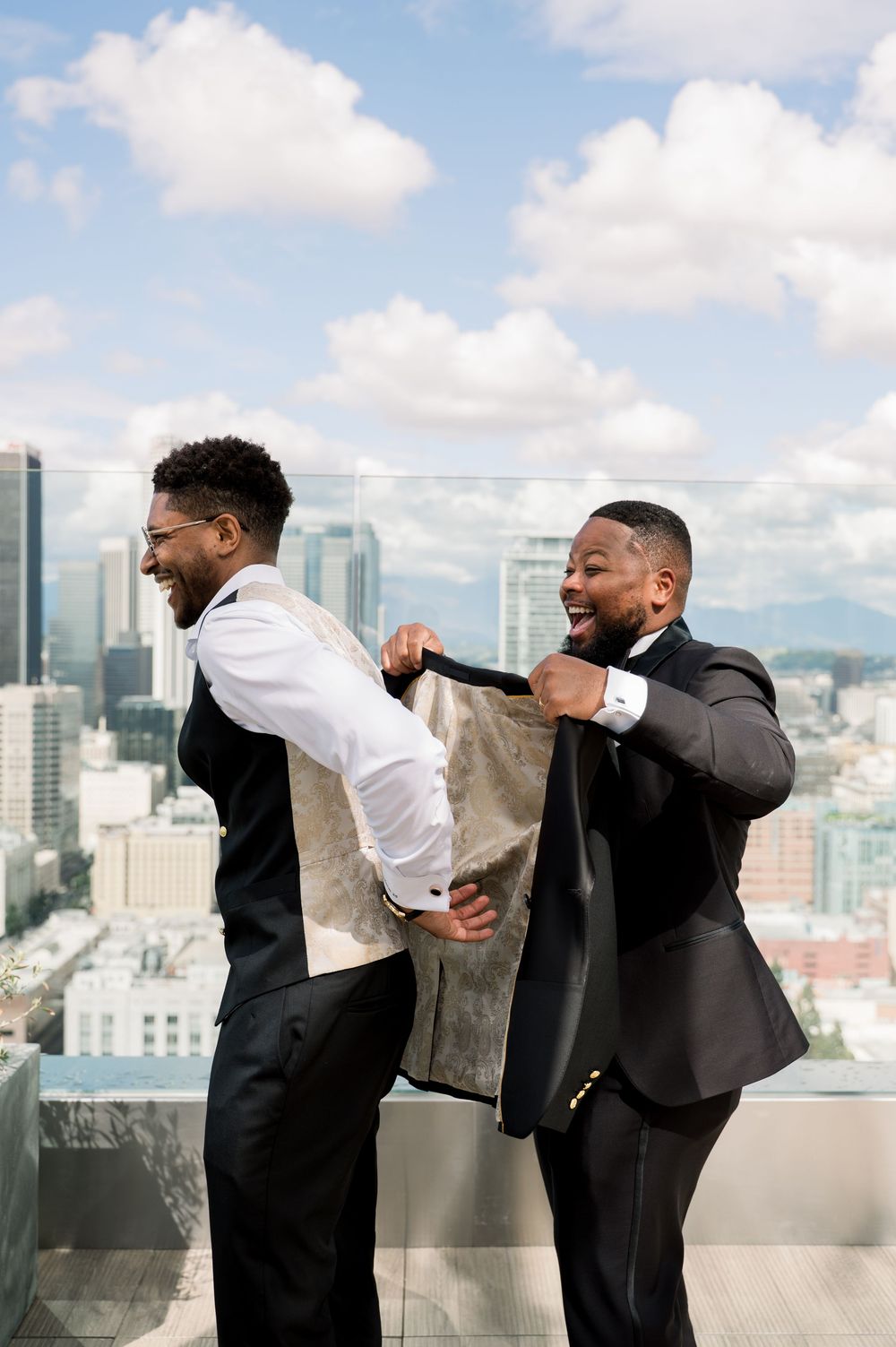 Two men in black suits laugh and share a fun moment on a rooftop terrace overlooking a city skyline on a sunny day.