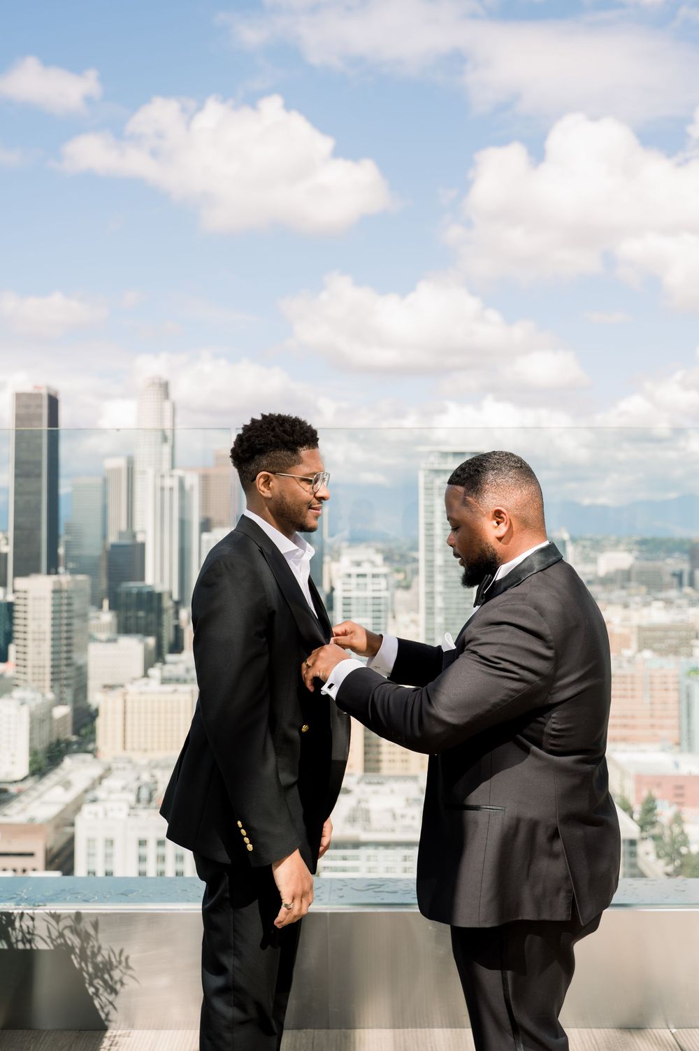 Two men in dark suits share an emotional moment on a rooftop terrace with a cityscape of high-rise buildings behind them.