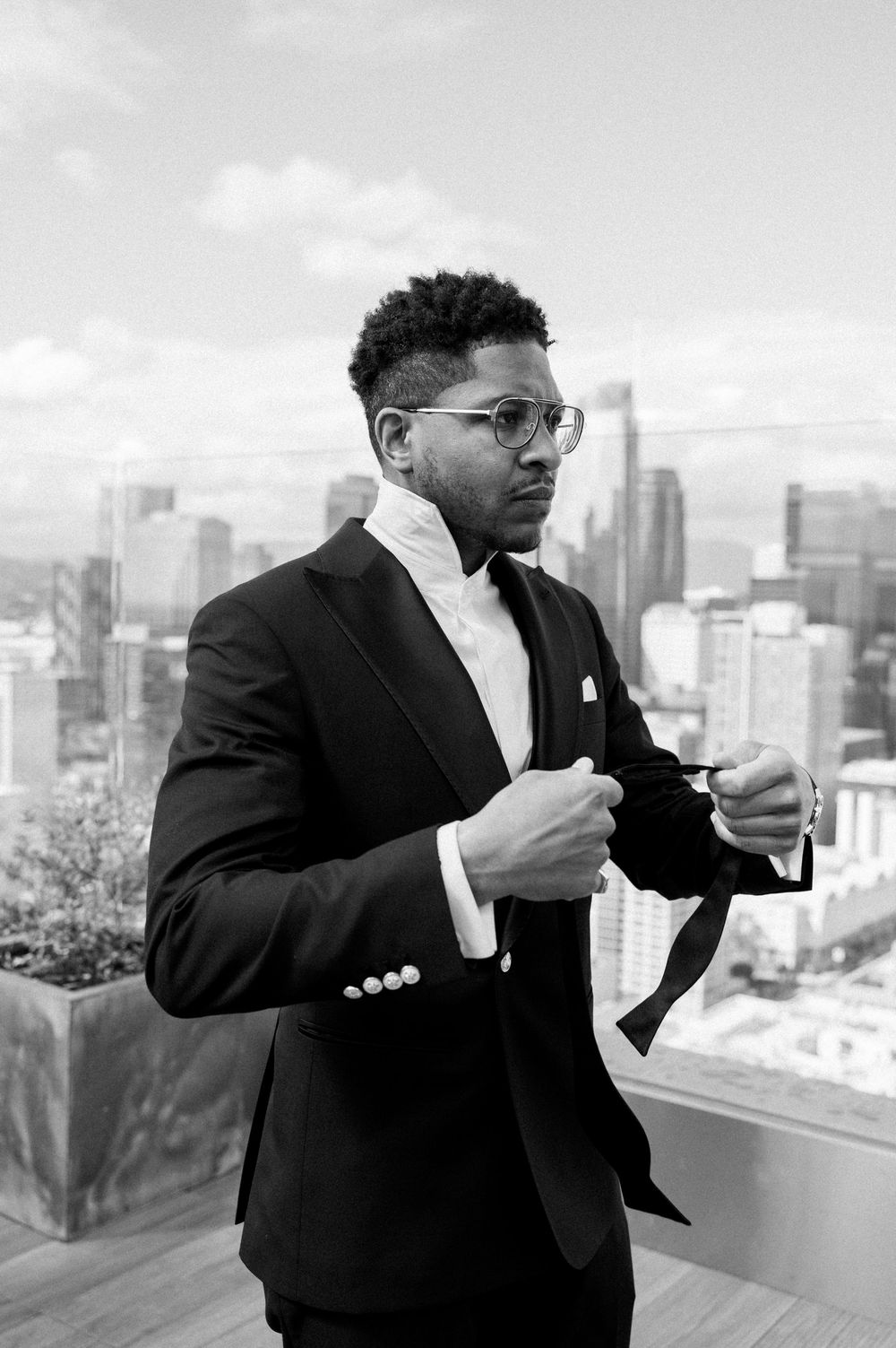 Professional black and white portrait of well-dressed figure adjusting tie on rooftop terrace with city skyline backdrop.