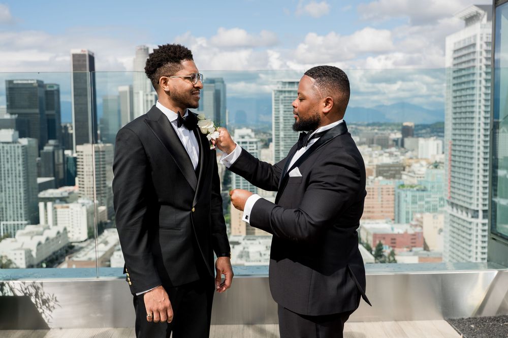 Two men in black suits and bowties share a laugh on a rooftop terrace overlooking a city skyline with skyscrapers.