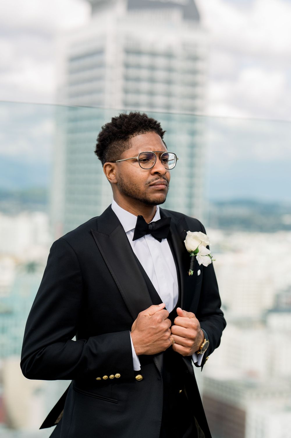 A groom in a classic black tuxedo and bow tie with a white boutonniere poses against a city backdrop.