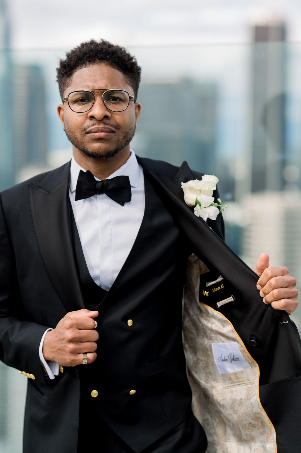 A groom in a black tuxedo with bow tie and white boutonniere poses against a cityscape background.