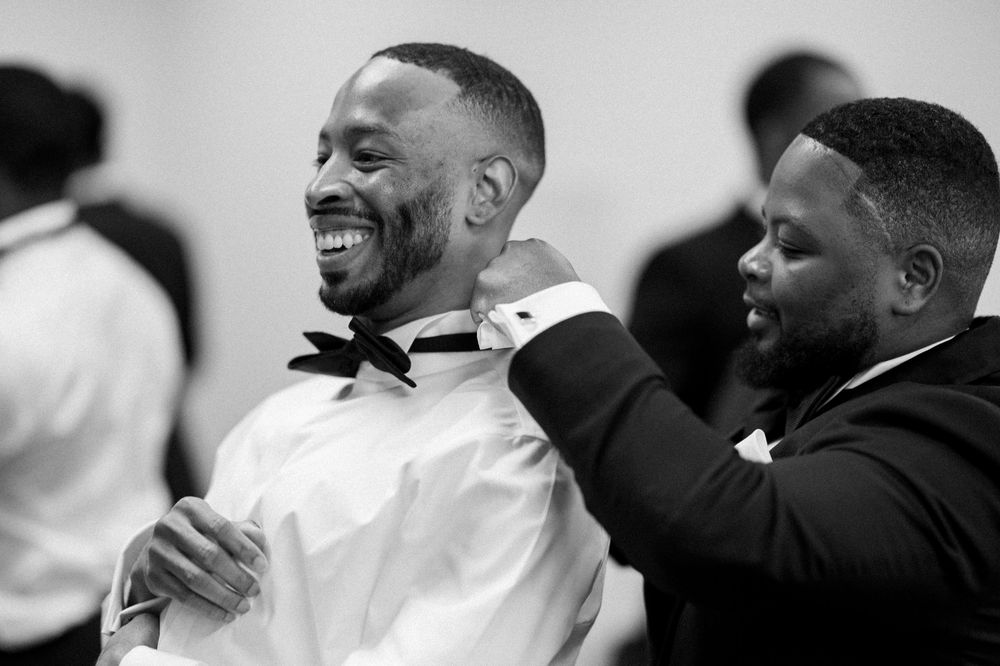 Black and white photo of two men laughing together, one adjusting the other's bowtie during formal preparation.